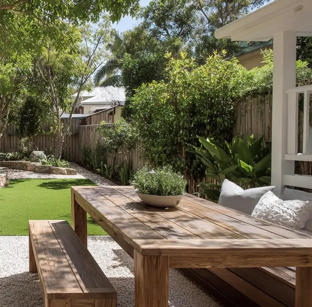 A rustic timber dining table and bench set sits on a white gravel area overlooking a lush OneGrass synthetic turf lawn