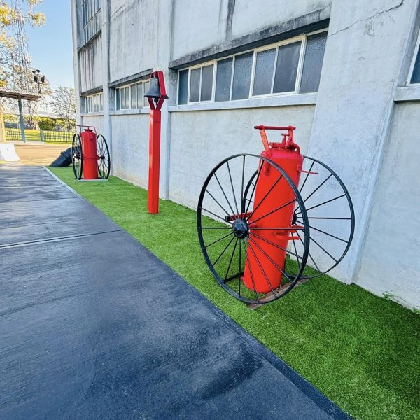 OneGrass synthetic turf strip featuring vintage red fire extinguishers and a bell outside an industrial building