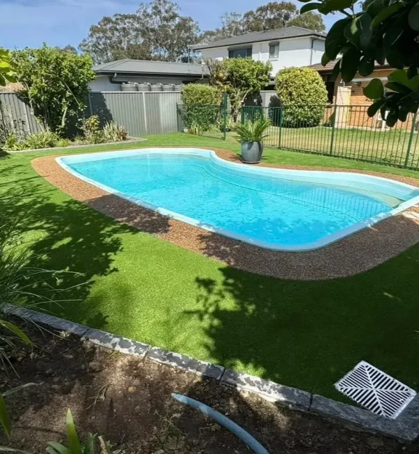 OneGrass synthetic turf provides a clean, green border for a backyard pool with pebble edging and garden beds