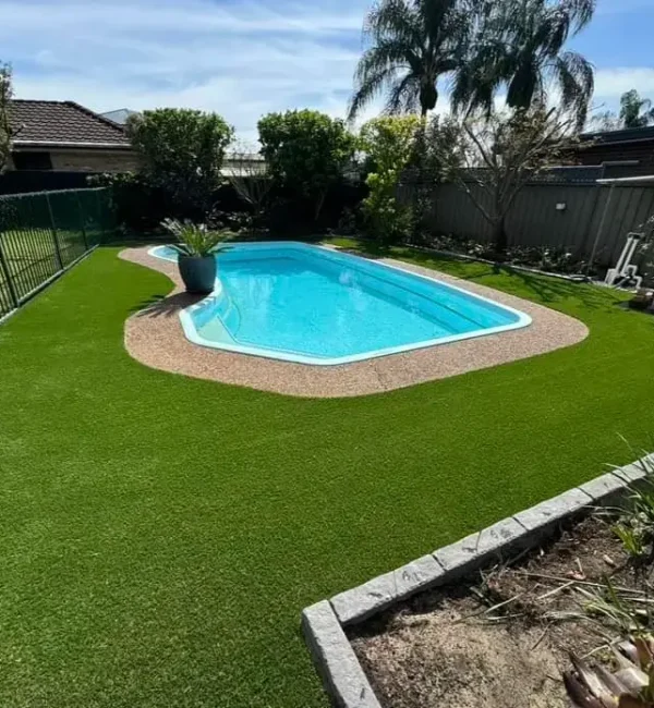 OneGrass synthetic turf enhances this backyard pool area, bordered by a dark stone garden bed and palm trees