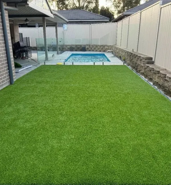 A neat backyard lawn featuring OneGrass synthetic turf adjacent to a swimming pool and stone retaining wall