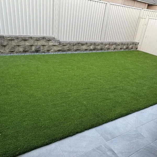 A perfectly level OneGrass synthetic turf lawn bordered by glass fencing, stone tiles, and a tiered retaining wall