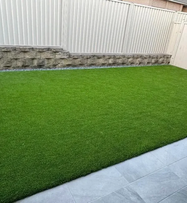 A perfectly level OneGrass synthetic turf lawn bordered by glass fencing, stone tiles, and a tiered retaining wall