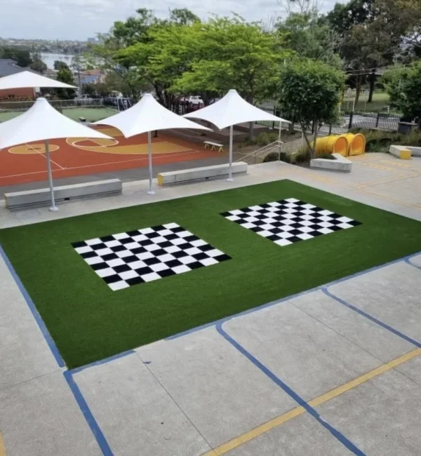 Two outdoor chessboard patterns integrated into a lush OneGrass synthetic turf area in a paved school courtyard