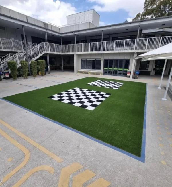 Two outdoor giant chessboard patterns integrated into a lush OneGrass synthetic turf area in a paved school courtyard