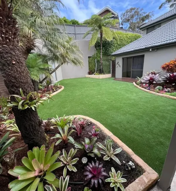 Lush OneGrass synthetic turf creates a pristine backyard lawn surrounded by stone-edged tropical garden beds