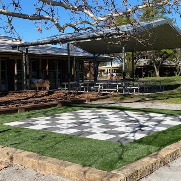 OneGrass synthetic turf borders a giant outdoor chessboard in a paved school courtyard area under a large canopy