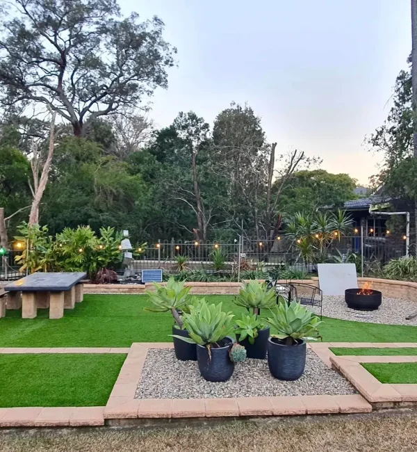 Modern backyard entertaining area featuring OneGrass synthetic turf, a fire pit, a stone table, and agave planters