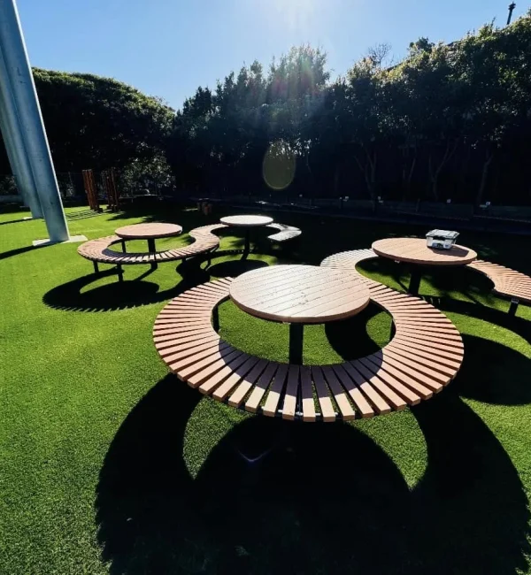 Circular timber tables sit atop a lush OneGrass synthetic turf surface in a sunlit school courtyard