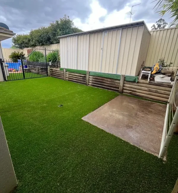 OneGrass synthetic turf creates a tidy backyard lawn beside a garden shed, timber retaining wall, and concrete pad