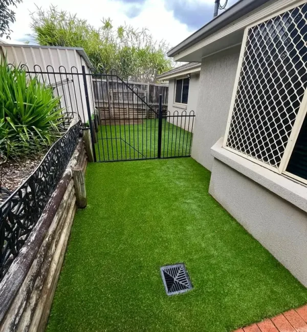 OneGrass synthetic turf installed in a narrow side yard with a drainage grate, black metal fence, and timber retaining wall