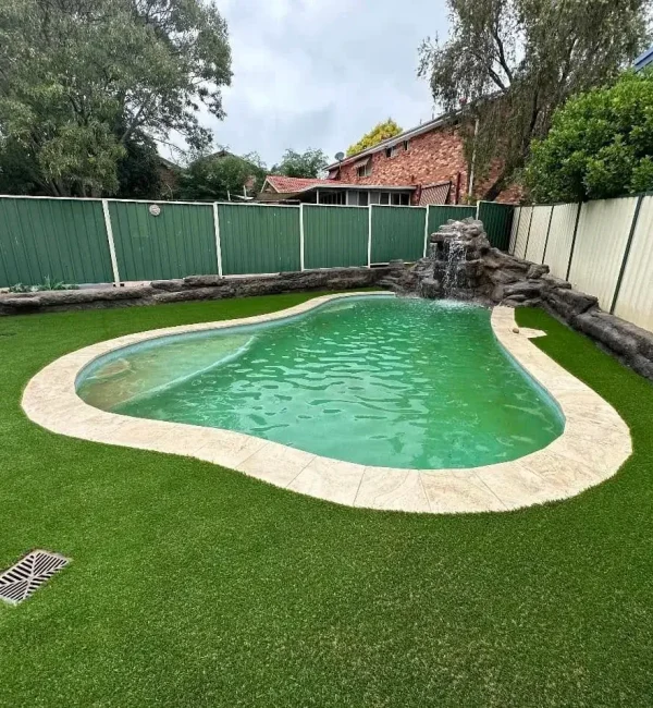 OneGrass lush artificial turf surrounds a backyard swimming pool with a rock waterfall feature and green metal fencing