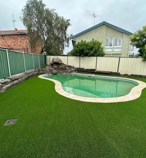 A spacious backyard featuring OneGrass artificial grass surrounding a curved swimming pool with rock features and fencing